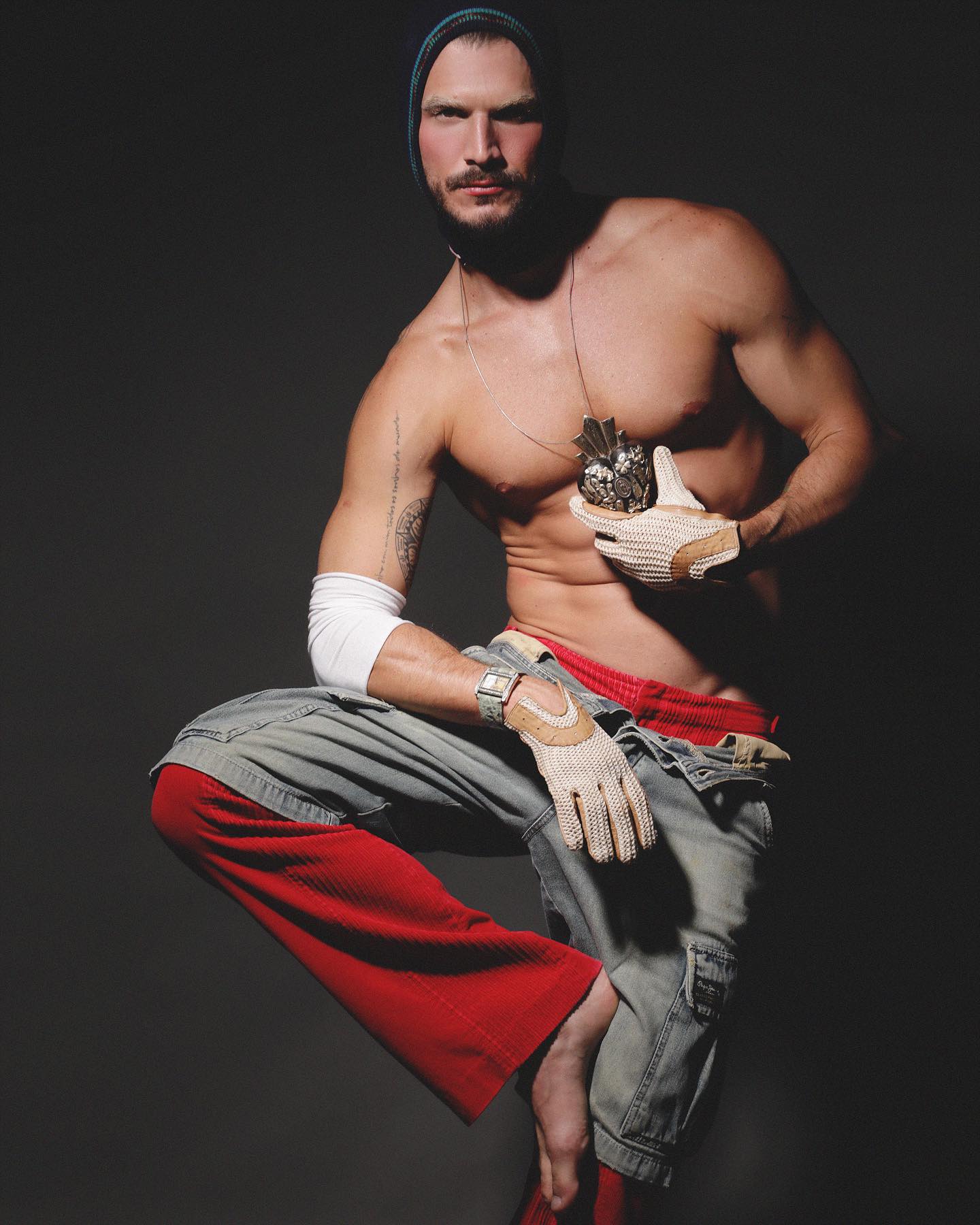 Alex Trevelin sentado com luvas, denim e calça vermelha em editorial de styling