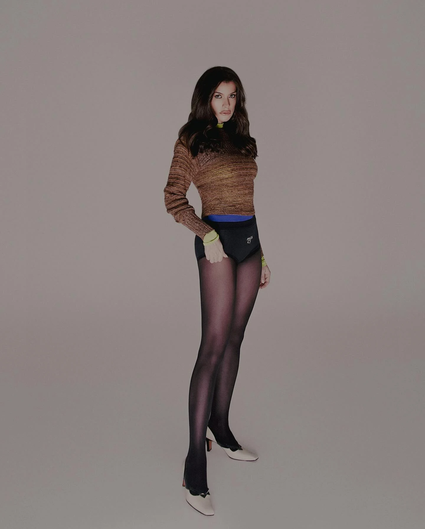Alinne Moraes em look com tricô marrom, hot pant preta, meia-calça e sapatos brancos
