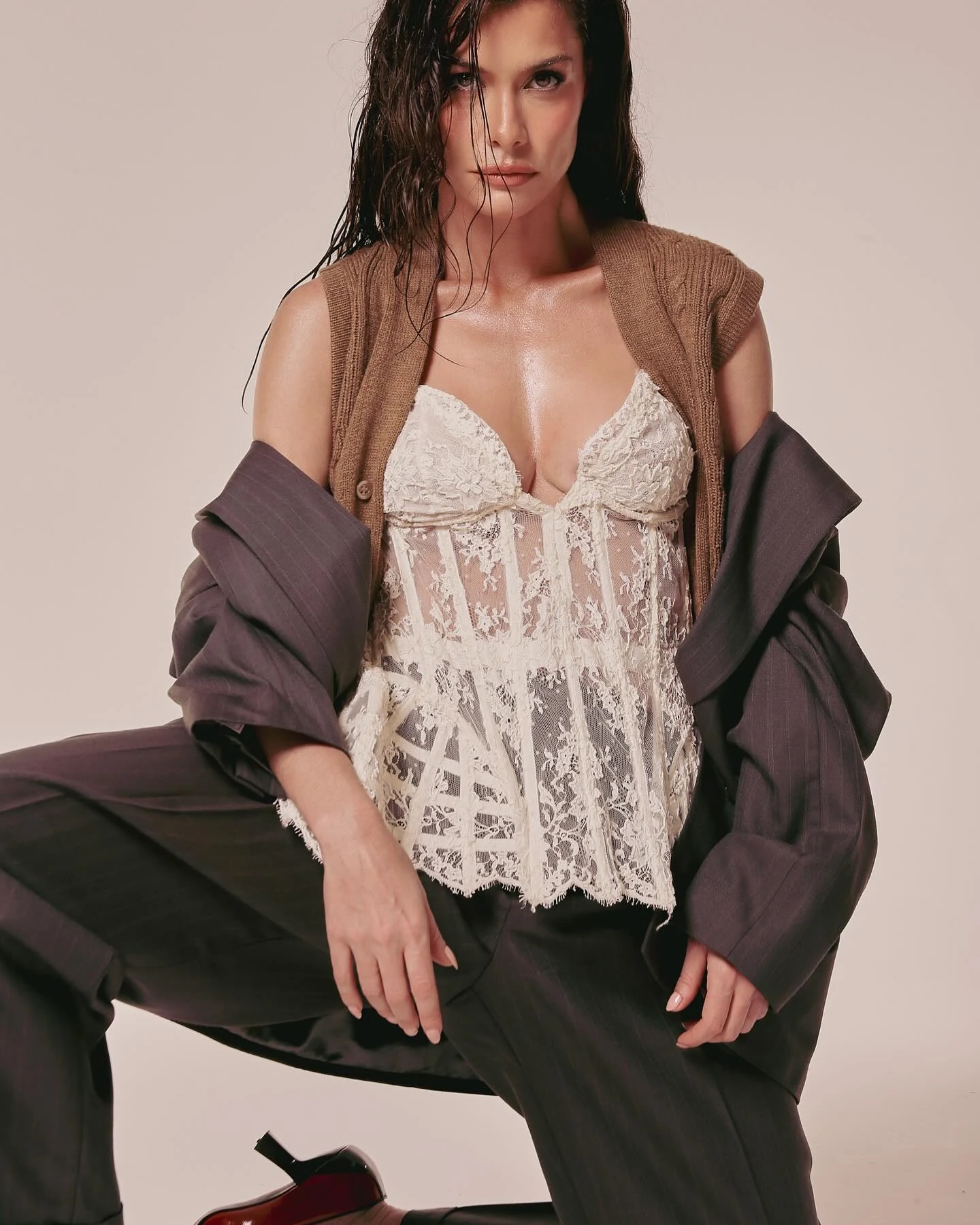 Alinne Moraes com corset de renda off-white, tricô marrom e blazer cinza em pose sentada