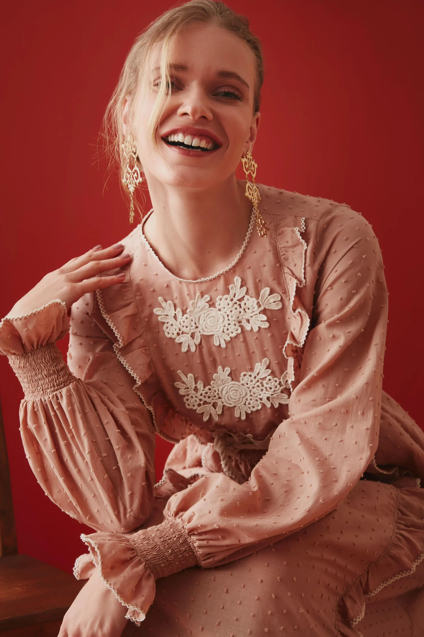 Retrato de Thais Custodio em vestido rosado bordado diante de parede vermelha